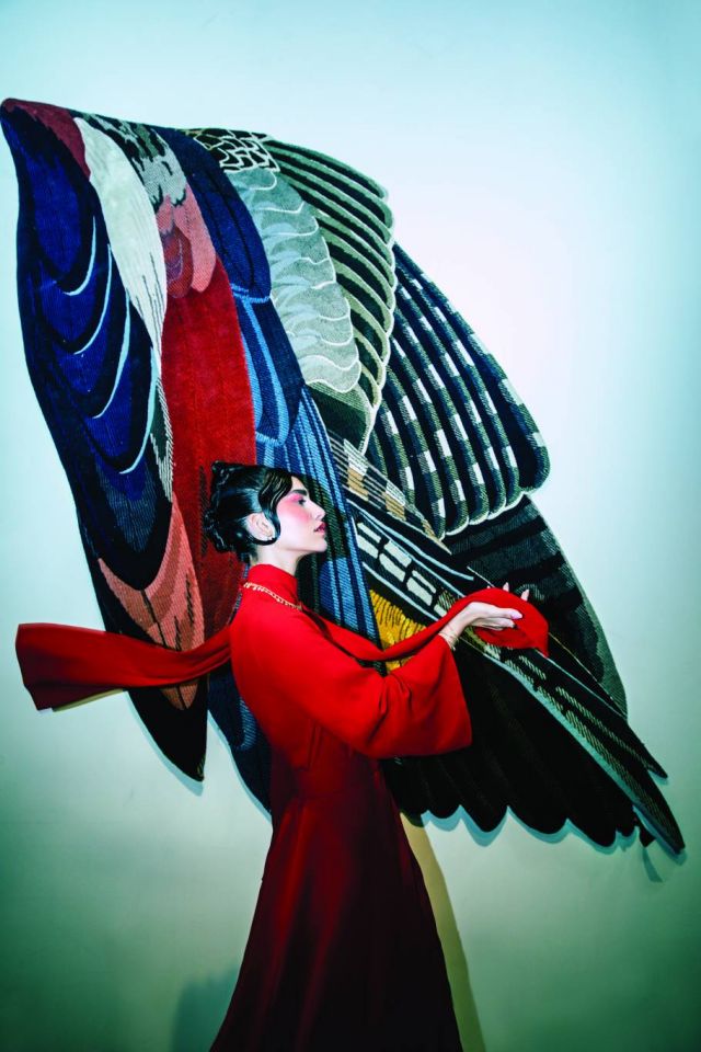 فستان من Carolina Herrera. أقراط للأذنين وعقد وخاتم وسوار من Boucheron.  بساط من الريش من تصميم  Maarten De Ceulaer محبوك يدوياً في النيبال باستعمال صوف الهيمالايا والحرير الطبيعي متوافرة لدى محلات إيوان مكتبي (السركال دبي)