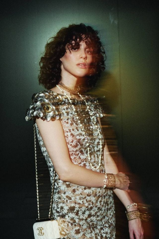 فستان من القطن المخرم باللون العاجي والأسود والوردي والأزرق مزين بأزرار مجوهرة وأزهار كاميليا عقد من المعدن والخرز الزجاجي والستراس. عقد من المعدن والجلد والستراس. أساور من المعدن والجلد والستراس. أساور من المعدن والخرز الزجاجي والستراس. حقيبة من الجلد الأبيض والخرز والمعدن. الكل من شانيل CHANEL